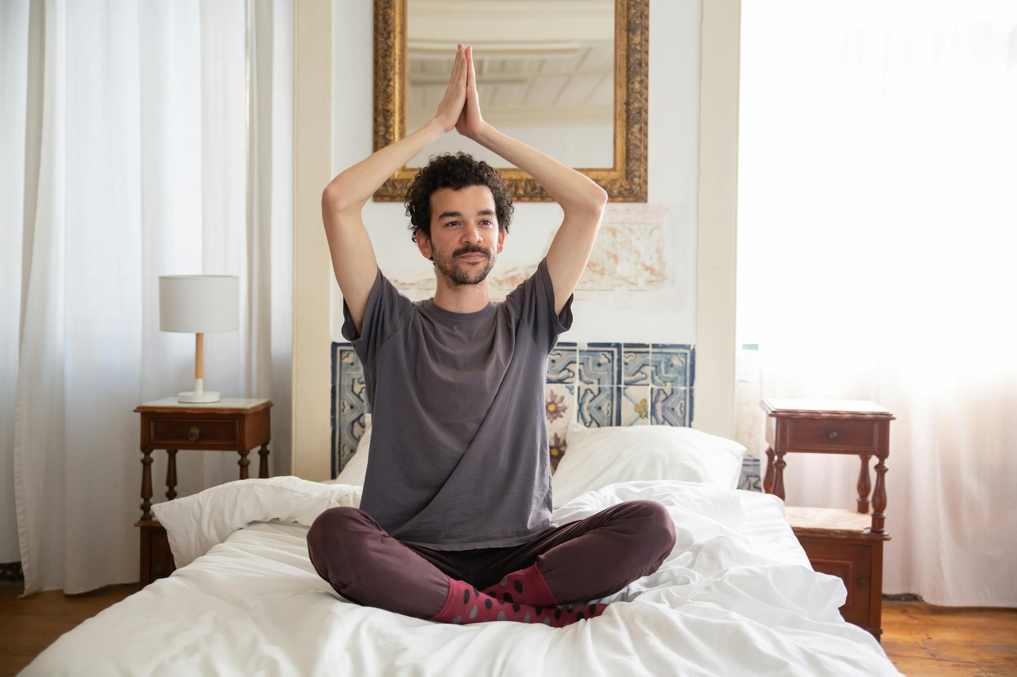 A man sits cross-legged on a bed, meditating with hands raised, in a sunlit bedroom.